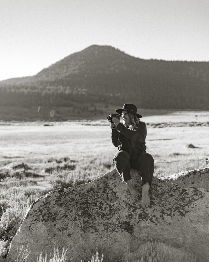 Lake Tahoe Photographer, sitting on a rock holding a camera, with mountains in the background.