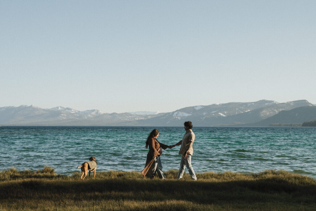 Couple and their dog walking along the waters edge in Lake Tahoe, with Mountains in the distance.